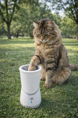 A fluffy brown tabby cat sits on park grass, reaching its paw into an Electric Pet Paw Washer with motorized silicone bristles. Trees and greenery form the background.