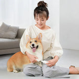 A woman sits on the floor by a gray sofa, using a Pawbby Pet Nail Grinder to groom her happy corgi’s paw. Both appear content in the bright, cozy room.