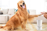 A happy golden retriever sits indoors with its paw raised to a person’s hand. An Electric Pet Paw Washer with motorized silicone bristles is in front of the dog, and a beige sofa can be seen in the background.